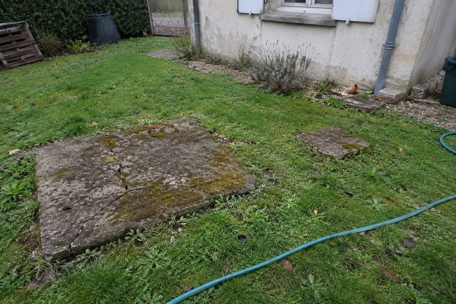 Deux anciens tampons en béton dans une pelouse, un grand et un petit, près du mur d’une maison.
